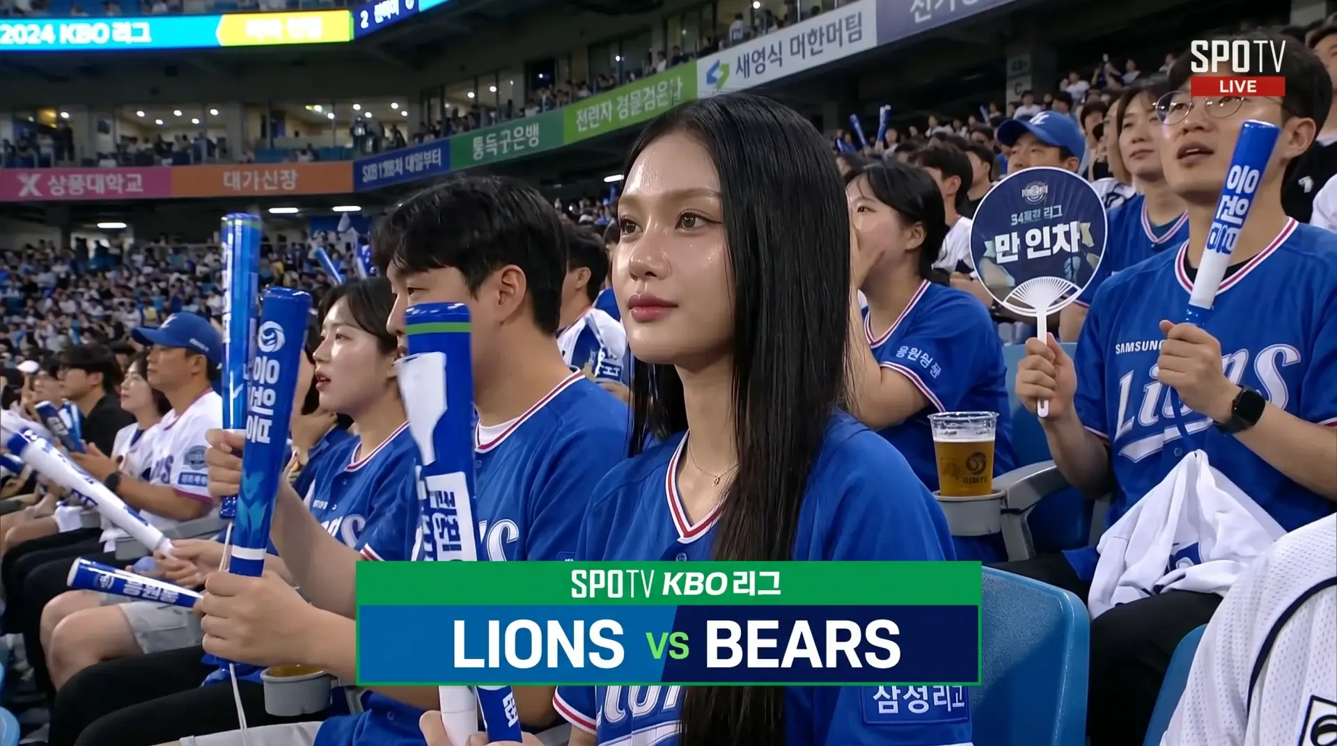 AI-generated KBO live broadcast still of a woman in a Samsung Lions jersey in a crowded baseball stadium crowd.